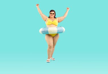 Mutlu, şişman, şişman, güneş gözlüklü, lastik yüzüklü, sarı mayo giyen ve elleri stüdyonun mavi arka planında olan şişman bir kadın. Yaz tatili ve tatil konsepti.