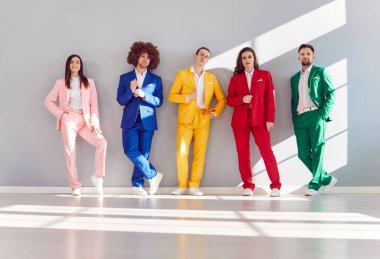 Renkli takım elbiseli bir grup pozitif genç gri bir duvarın önünde duruyor. Çeşitliliği ve takım çalışmasını temsil ediyorlar. Dinamik bir iş ekibinin bu portresinde mutlu bir şekilde gülümsüyorlar..