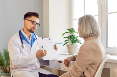 Erkek ciddi bir doktor yaşlı bir hastaya danışıyor, tıbbi danışma sırasında hastane ofisinde oturuyor. Hemşire, eski hastaya düzgün bir sağlık hizmeti sunuyor..