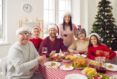 Mutlu yıllar ve mutlu Noeller. Noel Baba şapkalı, Noel Baba Festivali ev partisinde eğlenen çok nesilli bir ailenin grup fotoğrafı. Gülümseyen mutlu bir aile. Odadaki şenlik masasında birlikte poz veriyorlar..