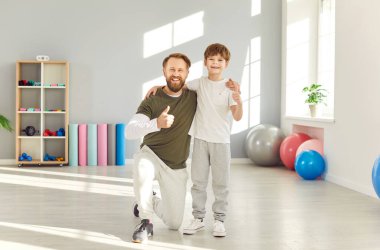 Spor kıyafetleri giymiş, spor giyinmiş oğluyla spor salonunda dikilen mutlu gülümseyen bir babanın portresi. Kameraya neşeli bir şekilde bakıyor ve birlikte antremandan sonra baş parmağını kaldırıyor. Aile spor konsepti.