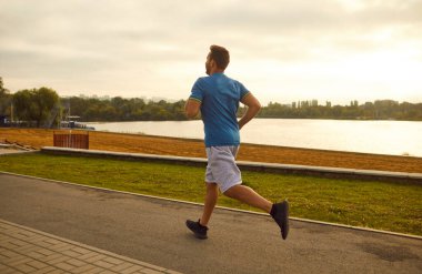 Nehir boyunca şehir parkında koşan atletik erkek koşucunun arka görüntüsü. Sağlıklı genç fitness adamı dışarıda koşuyor ve doğada egzersiz yapıyor. Sağlık ve sağlıklı yaşam tarzı kavramı.