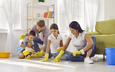 Mutlu gülümseyen genç ebeveynler ve iki kız ve erkek çocuk evdeki oturma odasında paçavra ve deterjanla birlikte yerleri temizliyorlar. Aile işleri, ev işleri ve ev işleri..