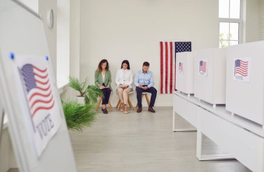 Oy verme merkezinde Amerikan bayrağıyla süslenmiş bir sıra beyaz oy kullanma kabiniyle oy kullanma merkezi. Amerikan seçmenleri sandık merkezinde oturuyorlar. Demokrasi ve seçim günü konsepti.