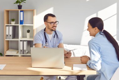 Kadın hastane ofisinde gülümseyen bir erkek doktorla ya da hemşireyle konuşuyor. Ziyaret sırasında konuşuyorlar, gülüyorlar ve tıbbi tavsiyelerde bulunuyorlar, olumlu ve destekleyici bir ortam yaratıyorlar..