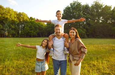 Mutlu aile taze yeşil parkta ya da şehir bahçesinde vakit geçirip doğanın tadını çıkarıyor ve birlikte eğleniyor. Anne, baba ve çocukların portresi yeşil çimlerde duruyor, arka planda güzel ağaçlar var.