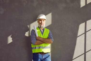 Kıdemli müteahhit, ustabaşı, inşaat mühendisi ya da mimarın portresi. Üniformalı, yelekli, gömlekli, beyaz kasklı kağıt rulo tutan ve gri duvarın yanında duran olgun sakallı adam.