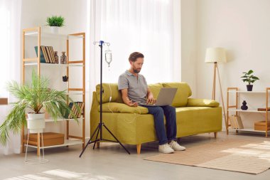 Genç adam kanında serum ve vitamin terapisi görürken evdeki oturma odasında dizüstü bilgisayarla oturuyor. Erkek kişi enjeksiyon tedavisi görüyor..