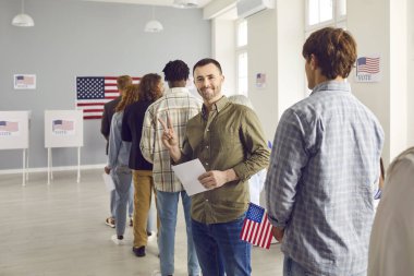 Mutlu Amerikalı seçmenler seçim günü oy kullanma merkezinde kayıt olurken zafer jesti yapıyorlar. Oy verme merkezinde kuyrukta bekleyen gülümseyen adam kameraya neşeli bakıyor..