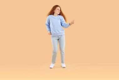 Neşeli genç kız neşeli bir şekilde eğleniyor ve ilham verici bir yüz ifadesiyle dans ediyor. Açık bej arka planda uzun dalgalı kızıl saçlı, sweatshirt, kot pantolon ve spor ayakkabı giyen bir kız..