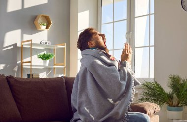 Hasta bir adamın evde peçeteye hapşırdığını gösteren yan görüntü. Evdeki kanepede oturan genç adam solunum hastalığından muzdarip mendille battaniyeye sarılmış. Mevsimsel enfeksiyon kavramı