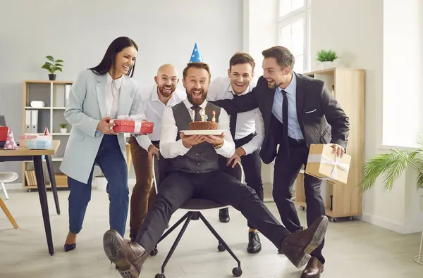Cheerful happy man celebrating his birthday in office with his colleagues. Man in party hat is sitting in office chair holding birthday cake while his satisfied colleagues surround him with gifts.
