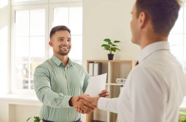 Mutlu gülümseyen adam, partneri ile ofiste durup başarıyı kutlarken ya da iyi bir anlaşma imzalarken el sıkışıyor. Genç ve neşeli erkek şirket çalışanları toplantı konusunda anlaşmaya vardılar..