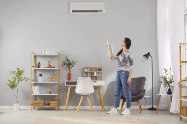 Kiracı ya da apartman sahibi modern klima sistemini evde değiştirir. Genç bir kadın güzel bir oturma odasında duruyor ve gri duvardaki klimanın ısısını ayarlıyor. AC kavramı