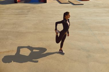 Spor elbiseli genç bir kadın şehir caddesinde spor yapıyor, gri beton spor sahasında duruyor, koşmaya hazırlanıyor ve bacak esnetme egzersizi yapıyor.
