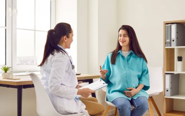 Doğum kliniğindeki doğum uzmanını ziyaret eden hamile bir kadın doktor masasında oturuyor. Jinekolog rapor tutuyor ve muayene sırasında danışmanlık yapıyor..