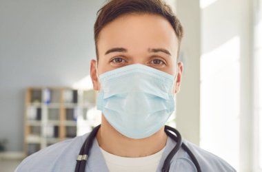 Tıbbi maske takmış, steteskoplu genç bir doktorun yakın plan portresi. Hastanedeki veya klinikteki hastalara yardım etmek için kameraya bakıyor. İlaç ve sağlık hizmetleri kavramı.