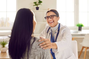 Mutlu doktor ve hasta eğleniyor. Beyaz ceketli, gözlüklü, genç bir kadınla konuşan, onu neşelendiren, elini omzuna koyan, gülümseyen, komik espriler yapan ve gülen bir kadın doktor.