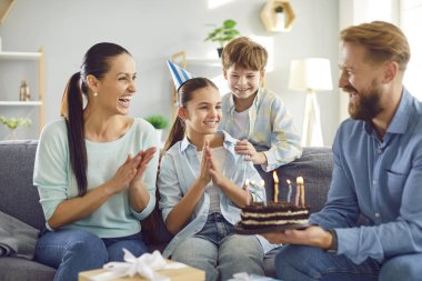 Mutlu gülümseyen, parti şapkalı ergen kız çocuğu evde anne, baba ve erkek kardeşle birlikte mumlarla pastaya bakarak dilek tutuyor. Mutlu aile ve bayram kutlamaları.