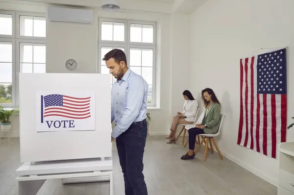 stock image Side view portrait of young american voter man standing at vote center with USA flags in voting booth making choice on paper ballot with people voters in background. Democracy and election day.