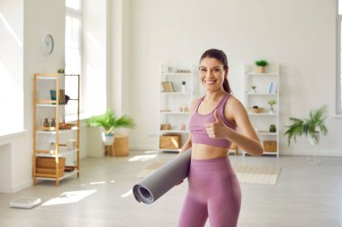 Leylak rengi spor sutyen ve spor pantolonu giymiş, elinde yoga paspası tutan, evdeki oturma odasında kameraya bakan, baş parmak hareketi yapan ve gülümseyen güzel bir genç kadın.