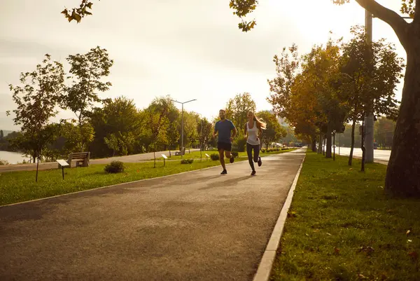 スポーツトレーニングをしている公園でジョギングスポーツウェアを着ている楽しい笑顔のスポーティカップル 若い男と女が屋外で走っている 自然の中でのワークアウト 健康的なライフスタイルのコンセプト — ストック写真