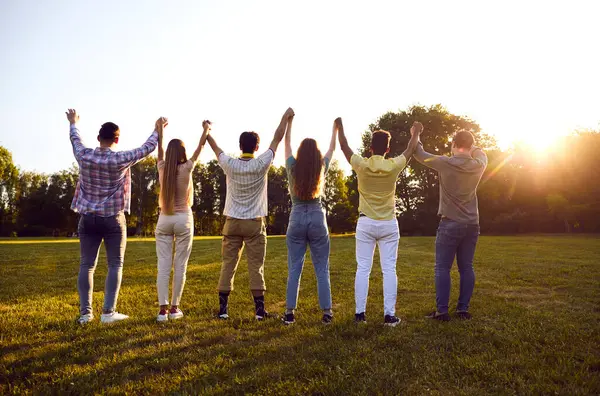Back view of a group of happy friends standing in a row holding hands with their hands up outdoors. Young men and women spending time together with raised arms enjoying sunset sky.