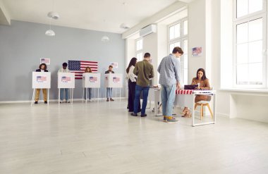 Bir grup gülümseyen Amerikan vatandaşı seçim günü oy kullanma merkezinde kayıt yaptırıyor ve oy pusulalarını oy verme kabinine koyuyorlar. Oy kullanma merkezinde sırada bekleyen seçmenler.