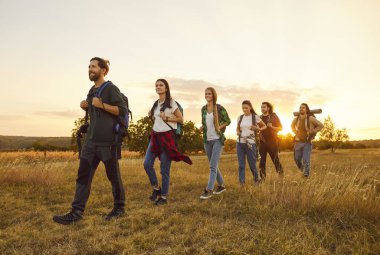 Şehir dışında yürüyüş yapan bir grup genç insan. Sırt çantalı mutlu erkek ve kadın turistler gün batımında kırsal bir alanda solgun çimlerde yürüyorlar. Seyahat, aktif tatil kavramı