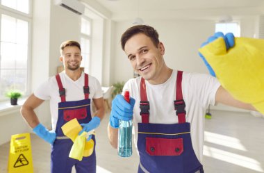Üniforma ve eldivenlerle süslenmiş pozitif temizlik ekibi neşeli bir portre için poz veriyor. Ellerinde kumaşla, ev idaresi uzmanlığı getiriyorlar, kendini işine adamış bir temizlik görevlisini somutlaştırıyorlar..
