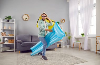 Şişme şilte ve bavul tutan neşeli komik adam evde oturmuş eğleniyor. Mutlu turistler yaz tatiline çıkıyorlar. Tatil kavramı.