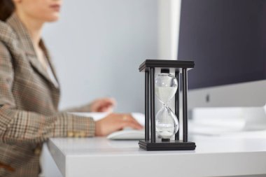 Teslim tarihine yetişmeye çalışan meşgul bir ofis çalışanının masasındaki kum saatini kapat. Masada oturan ve arka planda masaüstü bilgisayarı üzerinde çalışan bir kadınla yakın çekim. Zaman ve iş kavramı