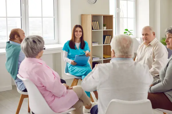 Group of elderly men and women sitting in a circle in nursing home and listening to young nurse during psychological therapy in session. Mental health and psychotherapy for senior people.