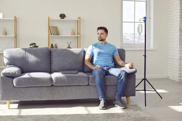 stock image Young man sitting on the couch at home or in clinic while receiving IV drip infusion and vitamin therapy in his blood. Attractive male person in casual clothes receiving injection therapy.