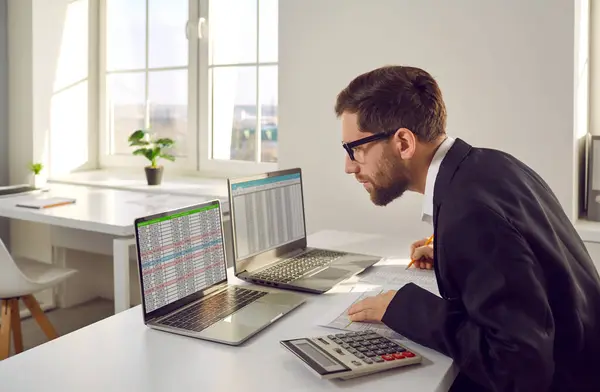在办公室的笔记本电脑上工作的企业会计 身穿西服 戴着眼镜的男人坐在办公桌前 用两台笔记本电脑 处理数据表 核对信息 — 图库照片