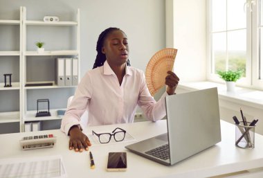 Genç Afro-Amerikan iş kadını. El yelpazesi kullanıyor. Isı ve yaz mevsiminin yüksek sıcaklıklarından mustarip. Ofiste sıcak hava dalgası sorunu.