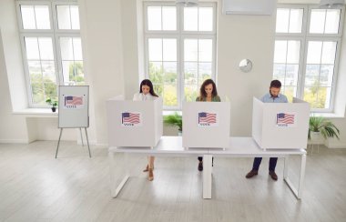 Kendine güvenen bir grup genç Amerikan vatandaşı oy verme merkezinde sırada bekliyor ve oy verme kabininde ABD bayrakları var. Seçim günü oy kullanma merkezindeki insanlar. Demokrasi kavramı.