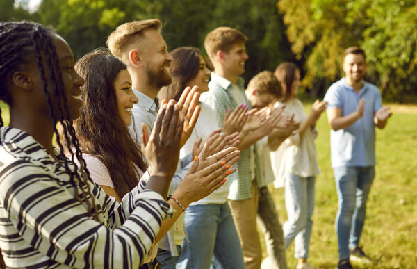 Счастливая группа друзей-студентов, стоящих в ряд и аплодирующих в летнем парке. Мультирасовые люди или коллеги улыбаются и хлопают в ладоши, наслаждаясь летом на культурных мероприятиях под открытым небом.