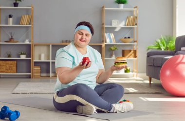 Genç, komik, şişman bir kadın halterli egzersiz yaptıktan sonra bir elinde hamburger, diğerinde elma tutarken neyi seçeceğini düşünüyor. Sağlıklı yaşam tarzı, diyet ve kilo verme kavramı.