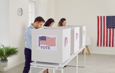 Yoğunlaşmış insanlar oy vermeden önce seçim pusulaları hakkında bilgi okuyorlar. Amerikan bayraklarıyla süslenmiş oy kullanma merkezindeki oy kullanma kabinlerinde genç erkekler ve kadınlar isimsiz oy kullanıyorlar..