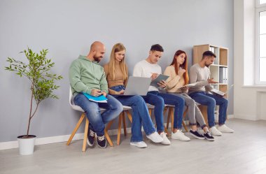 Koltuklarda oturan genç iş adamları ellerinde özgeçmişleri ve laptoplarıyla birbirleriyle sohbet ediyorlar. İş başvurusunda bulunanlar görüşme daveti bekliyorlar.