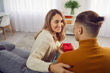 Elinde kırmızı hediye kutusu olan bir adam tatilde gülümseyen mutlu kadını tebrik ediyor. Evdeki koltukta oturuyor. Aşık mutlu bir çift. Dikkat, tebrikler ve sevgililer günü konsepti.