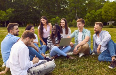 Mutlu, neşeli, çeşitli, çok ırklı, çok ırklı üniversite öğrencisi arkadaşlar dışarıdaki yeşil çimlerde oturuyor, akademik proje üzerinde çalışıyorlar, modern dizüstü bilgisayar kullanıyorlar ve birlikte eğleniyorlar.