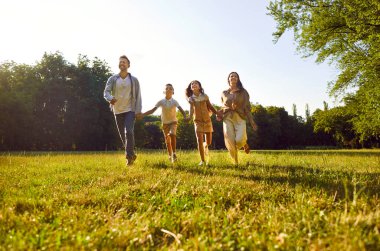 Mutlu aile, doğada boş vakitlerin tadını çıkarıyor. Neşeli anne, baba ve çocuklar el ele tutuşuyor ve güneşli bir günde güzel bir yaz parkında yeşil çimenlikte birlikte koşuyorlar.