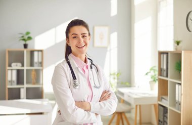 Ofisinde duran beyaz üniformalı esmer bir kadın doktorun portresi. Steteskoplu kendine güvenen kadın doktor kameraya neşeli görünüyor. Tıp konsepti.