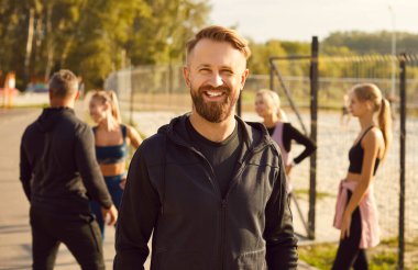 Şehir parkında spor egzersizleri yaptıktan sonra kameraya gülümseyen mutlu sakallı bir adamın portresi açık havada konuşan bir grup sporcuyla birlikte..