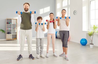 Aile dayanıklılık eğitimi. Neşeli, aktif ve çocuklu bir aile, parlak ve modern spor salonlarında dambılları birlikte kaldırıyor. Baba, anne, oğul ve kız spor eğitimi için hazır. Spor konsepti.
