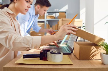 Lojistik ya da dağıtım merkezinde, depodaki depoda müşteriler için hazırlanan ve paketlenen dizüstü bilgisayarda çalışan bir adamla birlikte bayan yönetici. Envanter ve teslimat konsepti.