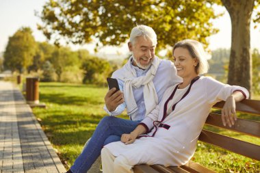 Halka açık bir parkta bankta oturan, birlikte cep telefonu kullanan ve dışarıda gülen mutlu kıdemli çift. Teknoloji ve emekliler konsepti.