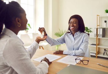 Profesyonel mutlu, gülümseyen Afro-Amerikan kadın emlakçı portresi ofiste oturan ve ev sahibi olmaya hazır bir kadına ev anahtarı veriyor. Emlak piyasası kavramı.
